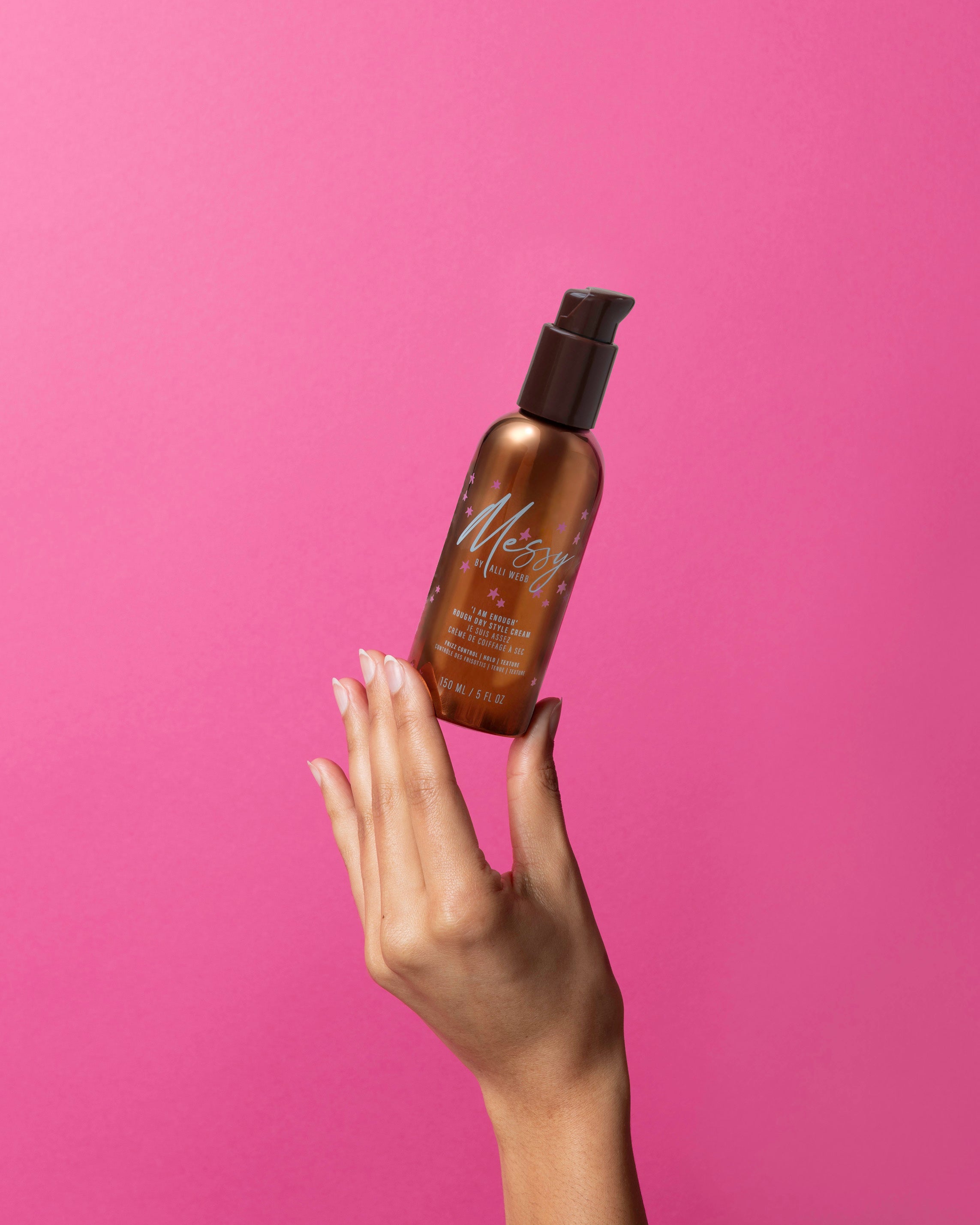 Hand holding the bronze 'I AM ENOUGH' Rough Dry Style Cream bottle against a bright pink background.